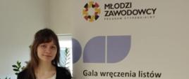 Zdjęcie: uśmiechnięta dziewczyna w biało-czarnym ubraniu stoi przy banerze programu stypendialnego :Młodzi zawodowcy"
