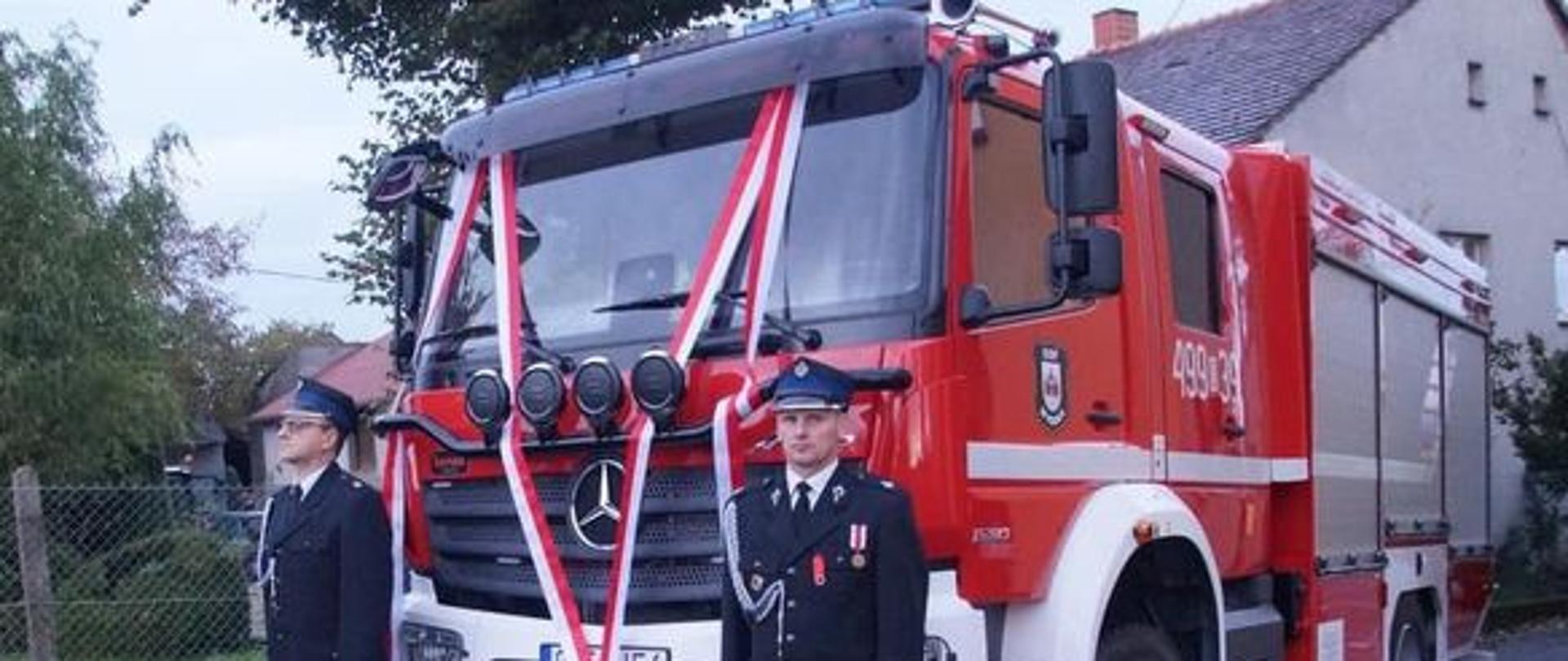 Na zdjęciu znajduje się samochód pożarniczy przystrojony w szarfy w barwach narodowych. Obok stoją strażacy ubrani w mundury wyjściowe