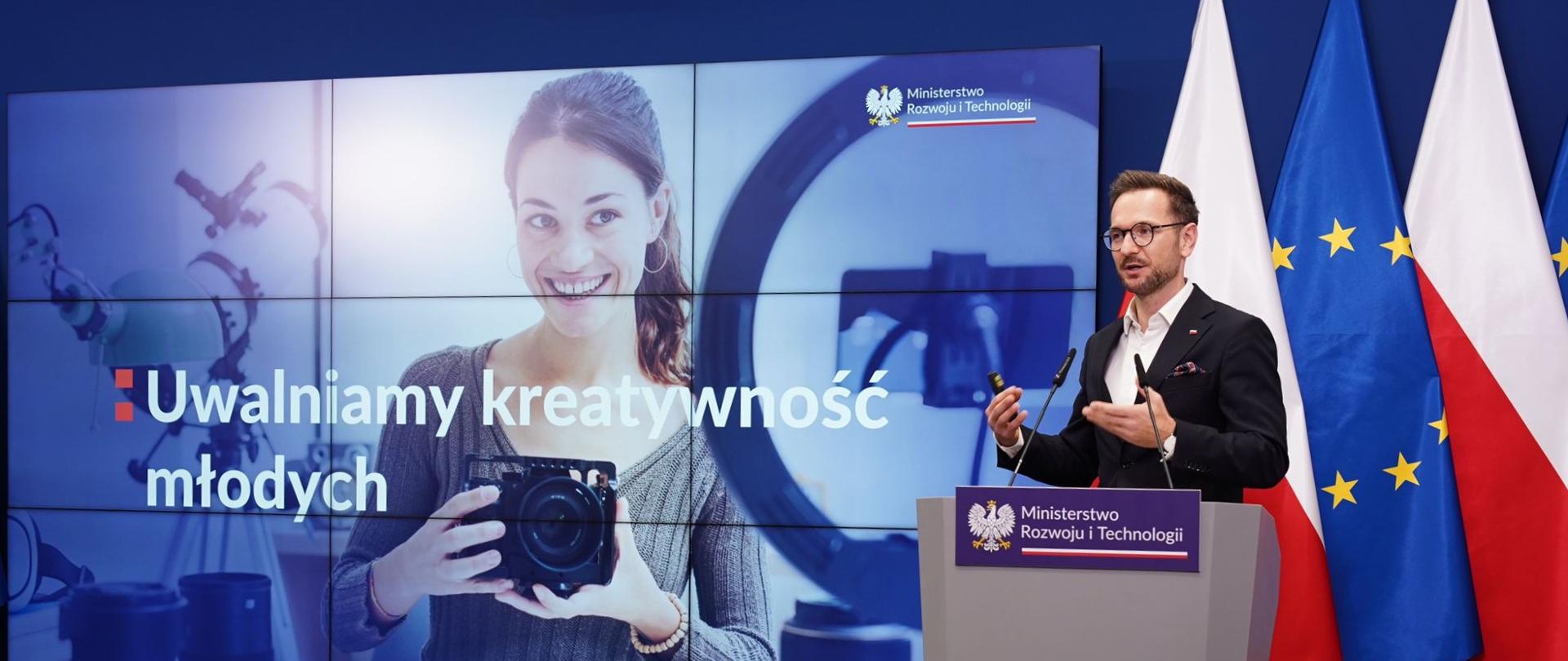 Konferencja ministra Waldemara Budy. Minister stoi po prawej stronie przy mównicy. Za nim flaga Polski i Unii Europejskiej. Za ministrem prezentacja. Na prezentacji dziewczyna, która trzyma aparat fotograficzny. Napis: Uwalniamy kreatywność młodych