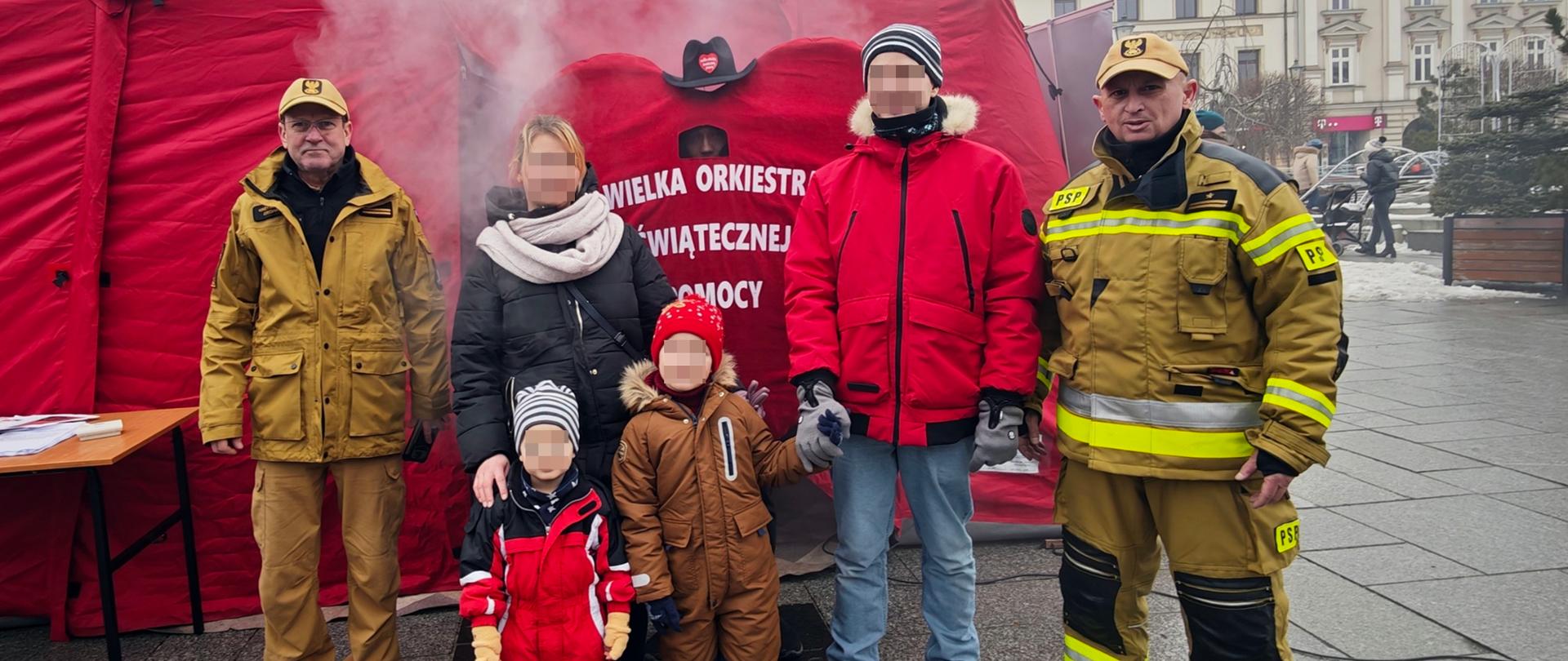 Na zdjęciu widać dwóch strażaków i czteroosobową rodzinę którzy stoją przed czerwonym namiotem. 