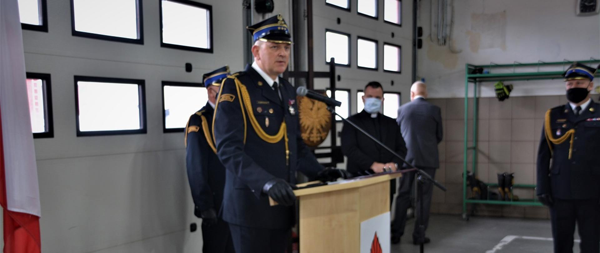 Na pierwszym planie w mundurze wyjściowym Zastępca Kujawsko-Pomorskiego Komendanta Wojewódzkiego Państwowej Straży Pożarnej przemawiający przy mównicy. Na zdjęciu widać również w mundurach wyjściowych Komendanta Powiatowego PSP w Tucholi i Dowódcę uroczystości oraz księdza kapelana wojewódzkiego strażaków. Wszyscy stoją w garażu JRG w Tucholi na tle bram garażowych. Na zdjęciu widać flagi państwowe i godło.