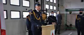 Na pierwszym planie w mundurze wyjściowym Zastępca Kujawsko-Pomorskiego Komendanta Wojewódzkiego Państwowej Straży Pożarnej przemawiający przy mównicy. Na zdjęciu widać również w mundurach wyjściowych Komendanta Powiatowego PSP w Tucholi i Dowódcę uroczystości oraz księdza kapelana wojewódzkiego strażaków. Wszyscy stoją w garażu JRG w Tucholi na tle bram garażowych. Na zdjęciu widać flagi państwowe i godło.