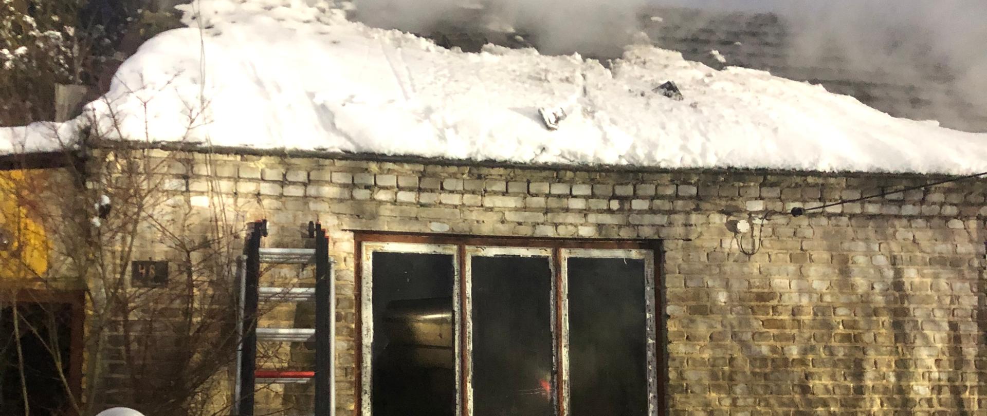 Strażak na pierwszym planie przy drabinie. Drabina jest oparta o budynek w którym doszło do pożaru. Dach budynku pokryty jest śniegiem. Z dachu wydobywa się dym. Zdjęcie w porze wieczornej.