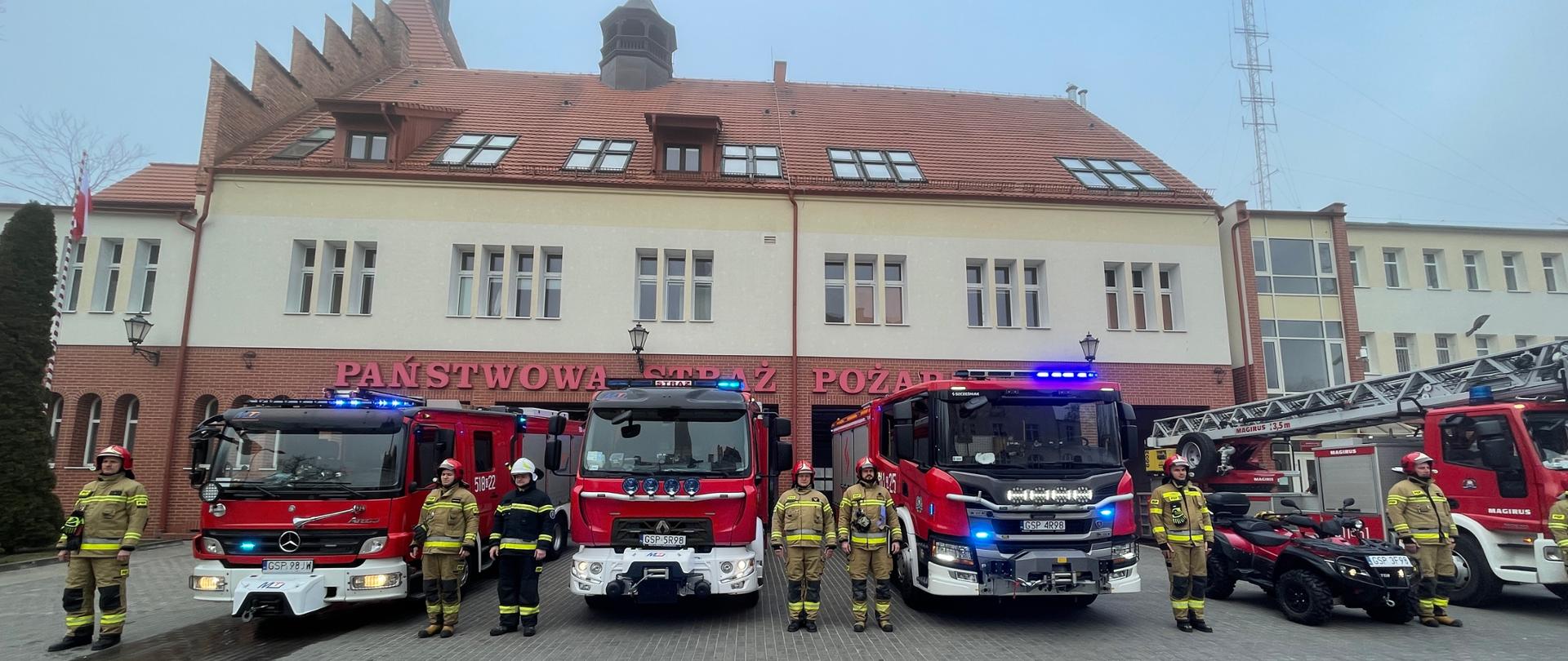 Przed budynkiem straży pożarnej stoją w rzędzie wszystkie samochody gaśnicze oraz quad i drabina mechaniczna. Obok nich stoją strażacy PSP i OSP ubrani w ubrania specjalne i hełmy. Na samochodach włączone są tylko sygnały świetlne