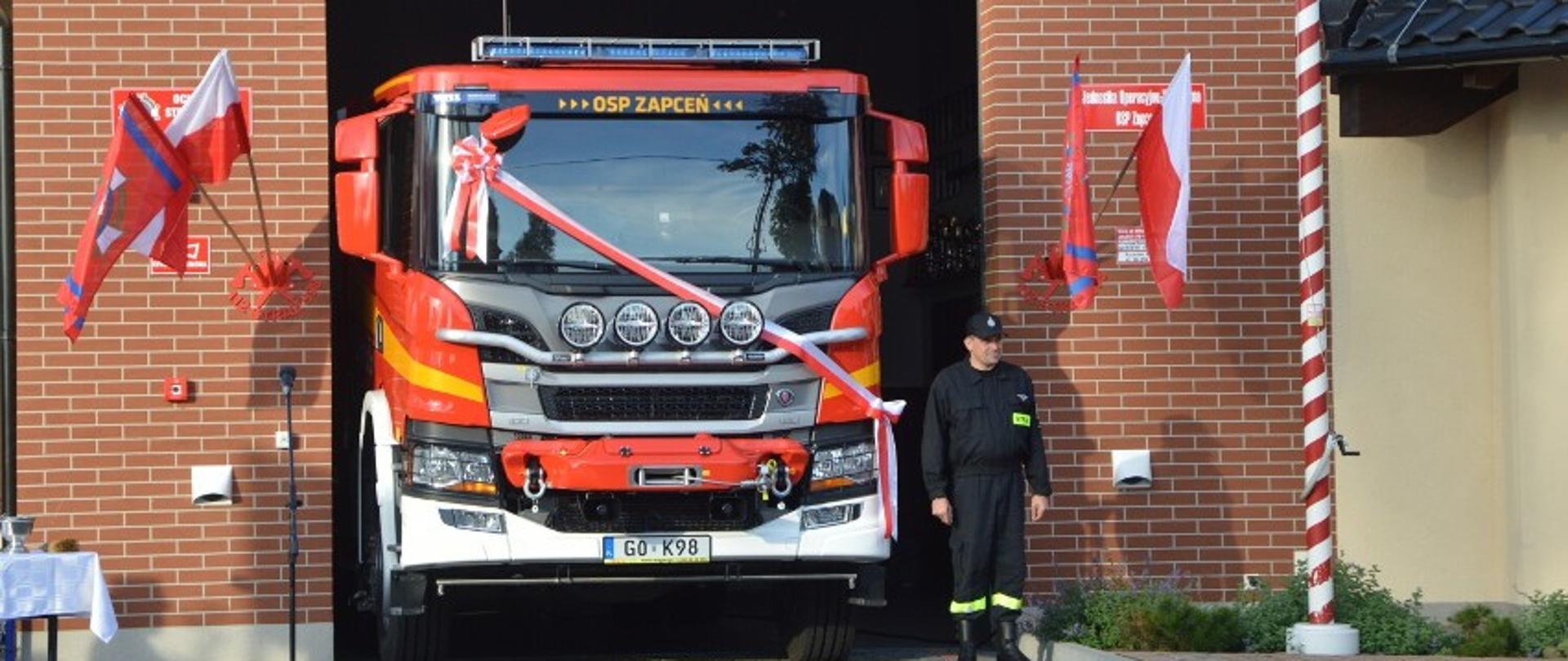 Zdjęcie przedstawia stojący samochód strażacki koloru czerwonego z przewieszoną biało-czerwoną wstęgą w bramie wjazdowej budynku, na którym zawieszone są flagi Polski i Związku OSP RP. Obok samochodu stoi strażak w mundurze koszarowym