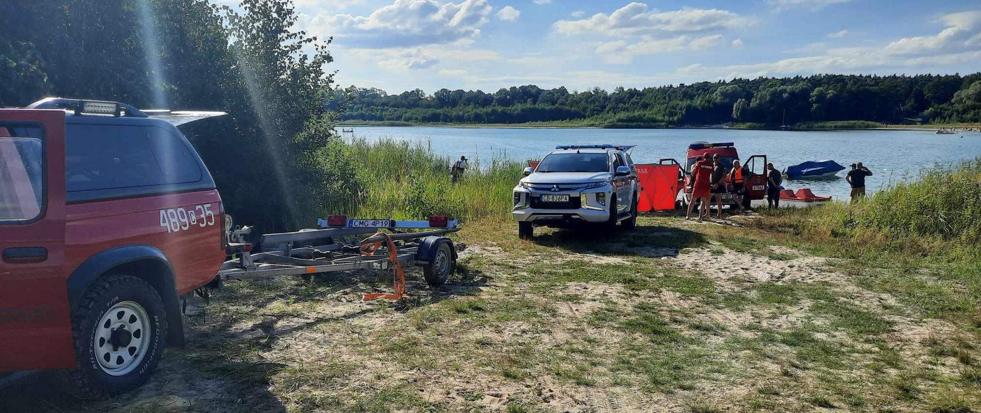 Na zdjęciu widać samochody straży pożarnej. Pierwszy samochód z lewej ma podczepioną przyczepkę. Z tyłu za srebrnym samochodem znajduje się parawan oraz kolejny samochód strażacki. Z tyłu widać jezioro i rozciągnięty za jeziorem las.