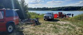 Na zdjęciu widać samochody straży pożarnej. Pierwszy samochód z lewej ma podczepioną przyczepkę. Z tyłu za srebrnym samochodem znajduje się parawan oraz kolejny samochód strażacki. Z tyłu widać jezioro i rozciągnięty za jeziorem las.