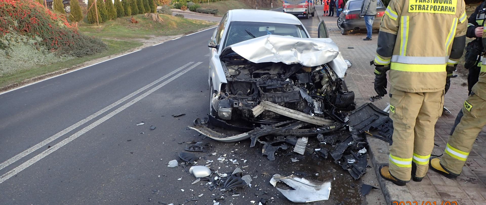 Zniszczony przód pojazdu po zdarzeniu z innym. Strażak OSP stojący plecami do kadru przed pojazdem
