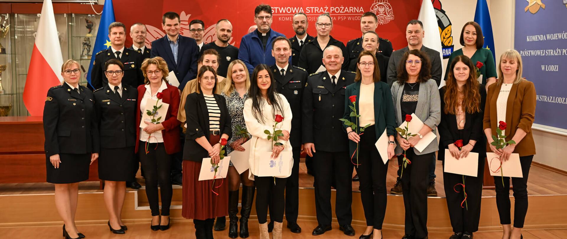 Zdjęcie grupowe uczestników uroczystości na tle czerwonej ścianki z godłem Polski i napisem „Państwowa Straż Pożarna”. Cywilne osoby trzymają róże i dokumenty, a towarzyszą im strażacy w mundurach galowych.