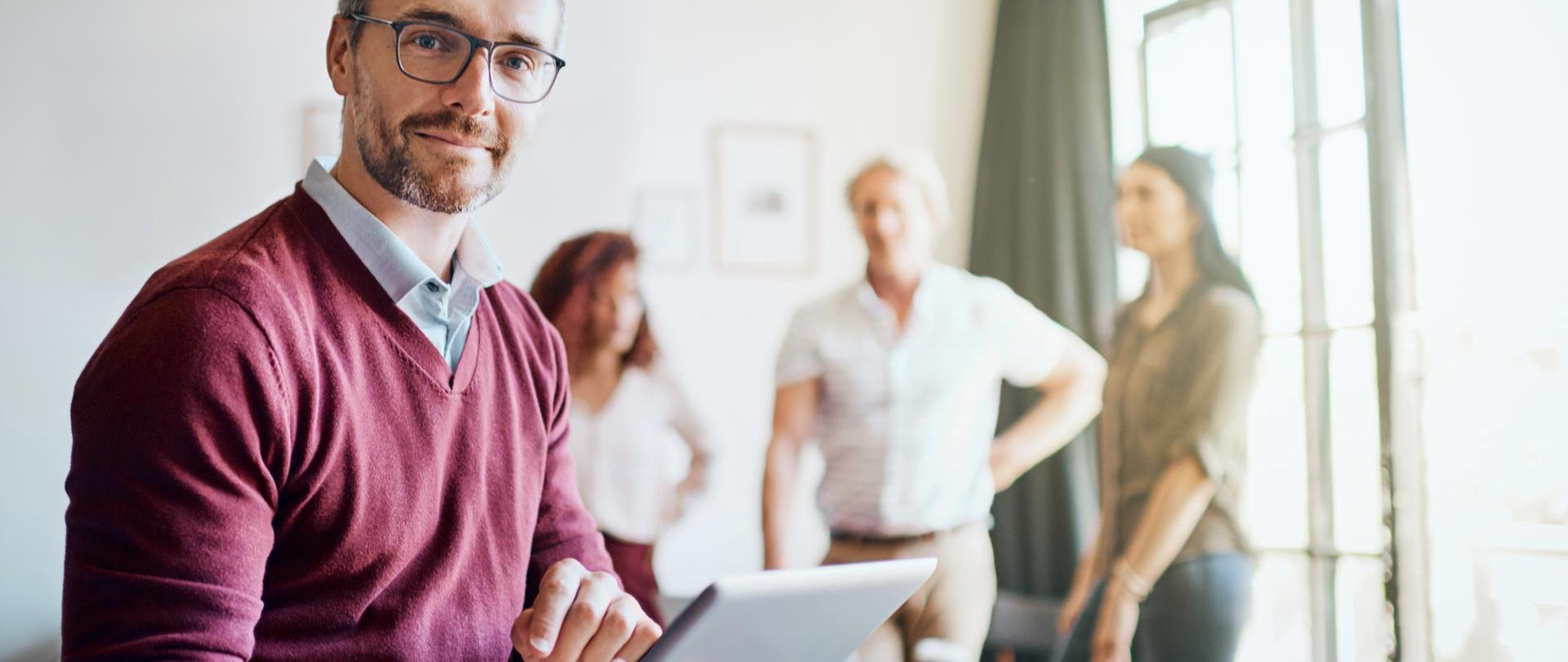 mężczyzna trzymający tablet, za nim rozmawiają stojąc wokół stołu dwie kobiety i mężczyzna