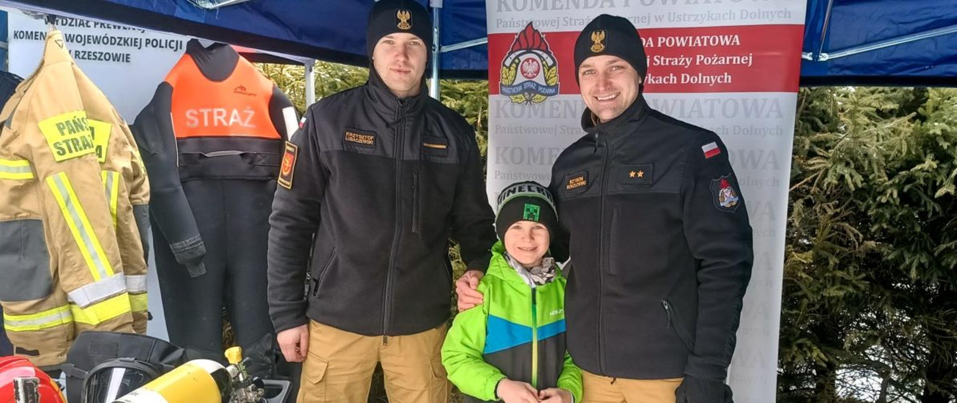 Zdjęcie przedstawia dwóch funkcjonariuszy Straży Pożarnej oraz chłopca z siatką i logiem psp na stoisku Państwowej Straży Pożarnej. Na stoisku widać sprzęt strażaków. 