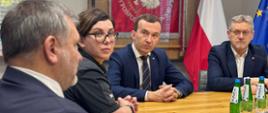 Oficjele siedzą przy stole, na pierwszym planie jest rozmazana postać, Minister na którą patrzy sie mężczyzna w garniturze, Wojewoda.