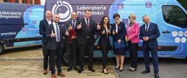 Grupa ludzi wśród których jest minister Czarnek stoi na dworze, pokazują uniesione w górę kciuki, za nimi niebiesko-fioletowa furgonetka z napisem Laboratoria przyszłości.