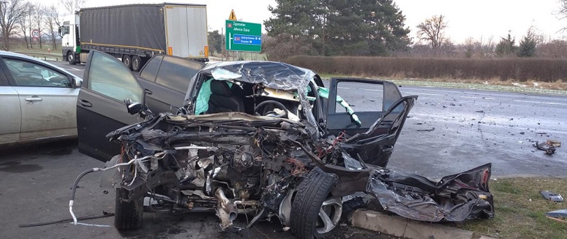 Widok od przodu na samochód osobowy, ciemny kolor karoserii. Przód samochodu całkowicie zniszczony. Brak silnika, skrzyni biegów oraz przedniej szyby. Pogięta pokrywa komory silnika leży obok samochodu Drzwi pojazdu otwarte. W tle zdjęcia stoi samochód ciężarowy z naczepą.