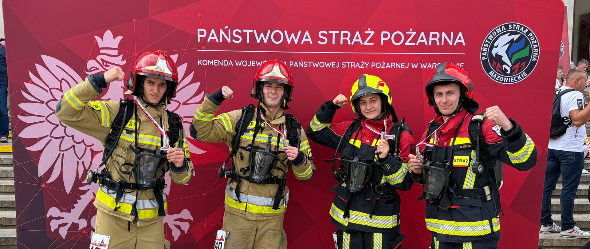 Drużyna strażaków z KP PSP w Czarnkowie pozuje na tle ścianki Komendy Wojewódzkiej PSP w Warszawie, prezentując medale i podnosząc zaciśnięte pięści w geście zwycięstwa.