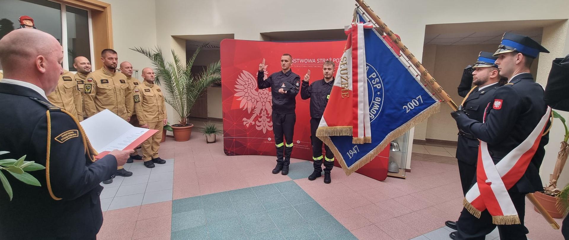 Dwóch nowych funkcjonariuszy Państwowej Straży Pożarnej składa ślubowanie podczas uroczystości w Komendzie Powiatowej PSP w Głogowie. Przed nimi stoi funkcjonariusz w mundurze galowym odczytujący rotę ślubowania, a obok znajduje się poczet sztandarowy. W tle stoją pozostali strażacy.