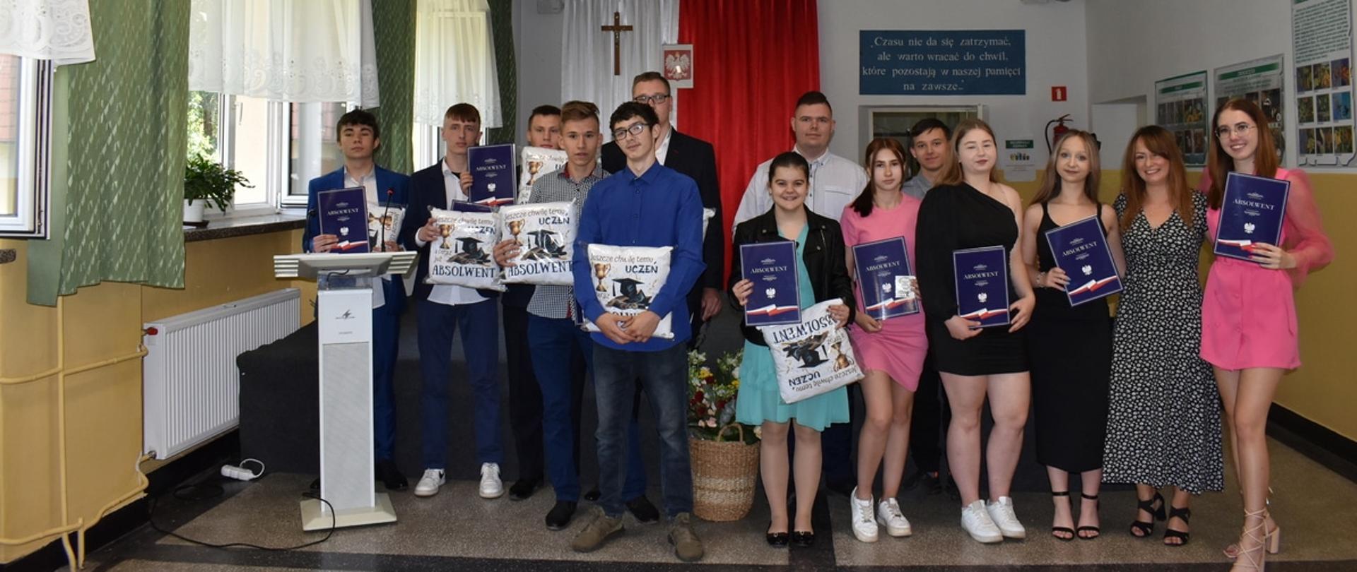 Pamiątkowe zdjęcie grupowe absolwentów oraz nauczycielki w sali szkolnej. Młodzi ludzie trzymają granatowe dyplomy z napisem „ABSOLWENT” oraz ozdobne białe poduszki. W tle widać biało-czerwoną dekorację z godłem i krzyżem.