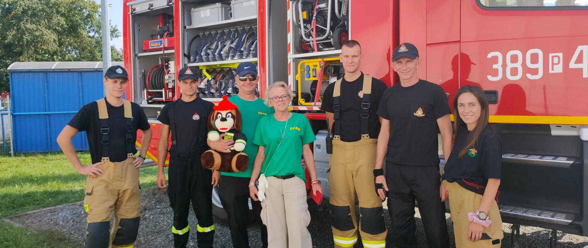 Strażacy oraz osoby cywilne stoją na tle samochodu strażackiego.