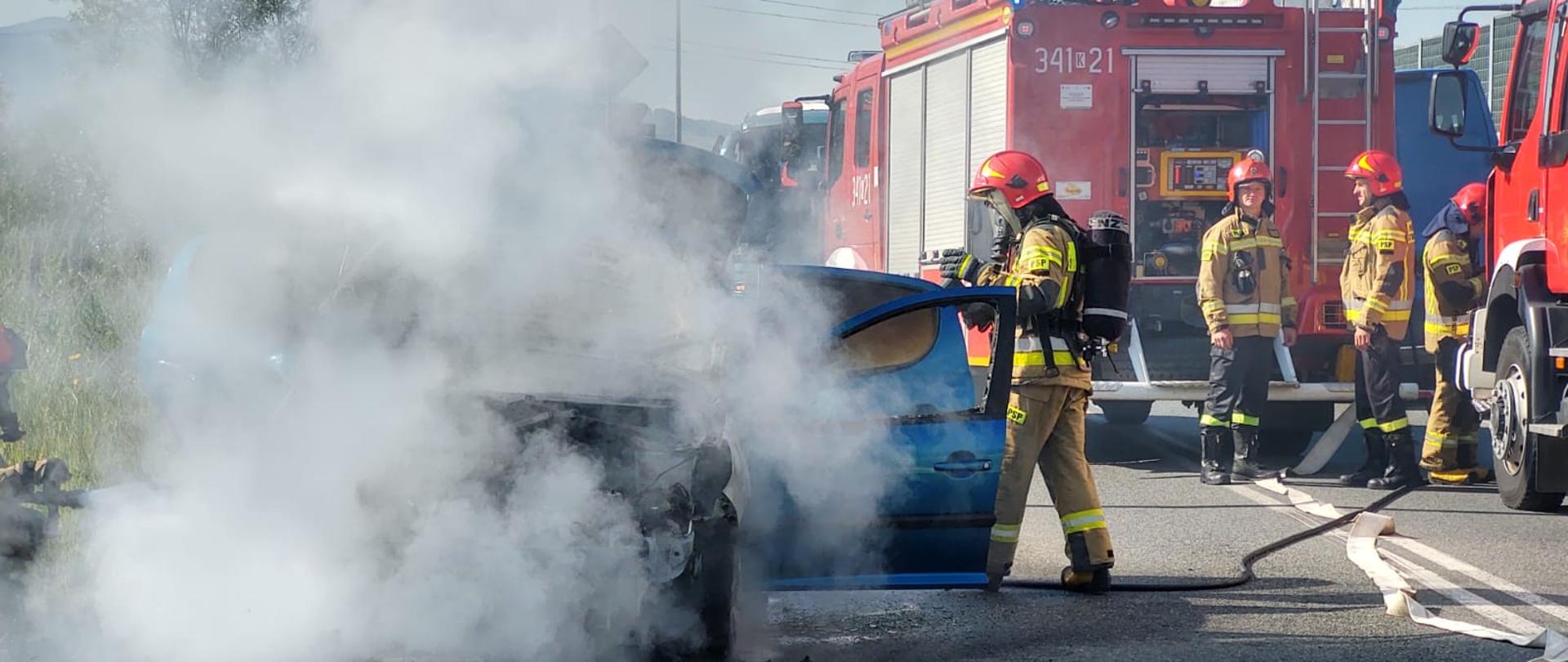 Strażacy gaszą pożar samochodu osobowego, z którego wydobywa się dym.