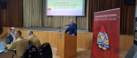 Strażacy ubrani w granatowe mundury galowe, oraz pisakowe mundury siedzą przy stolikach, w tyle rolap z logiem Państwowej Straży Pożarnej na tylnej ścianie kotary oraz ekran z wyświetloną prezentacją. Przy mównicy stoi starosta ubrany w garnitur, przed sobą ma mikrofon