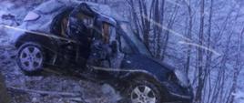 Na zdjęciu prószy śnieg; samochód ciemnego koloru poza jednią z widocznym rozbitym bokiem po uderzeniu w drzewo