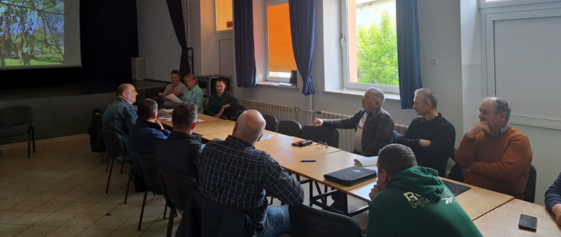 Grupa osób siedzi przy stole konferencyjnym w jasnej sali z dużymi oknami. W tle wyświetlana jest prezentacja przedstawiająca drzewo lub sad. Spotkanie ma charakter szkoleniowy lub informacyjny.