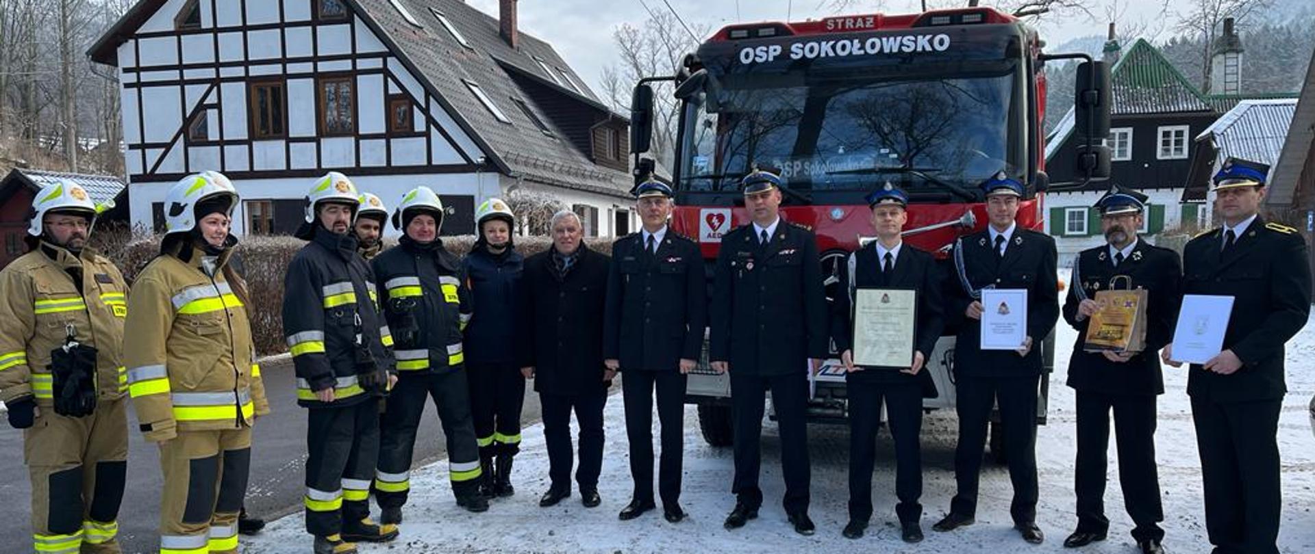 Zimową porą strażacy w mundurach bojowych w kolorze piaskowym i granatowym, oraz w granatowych mundurach wyjściowych. Część z nich w rękach trzyma dokumenty. W tle czerwony wóz strażacki oraz domy