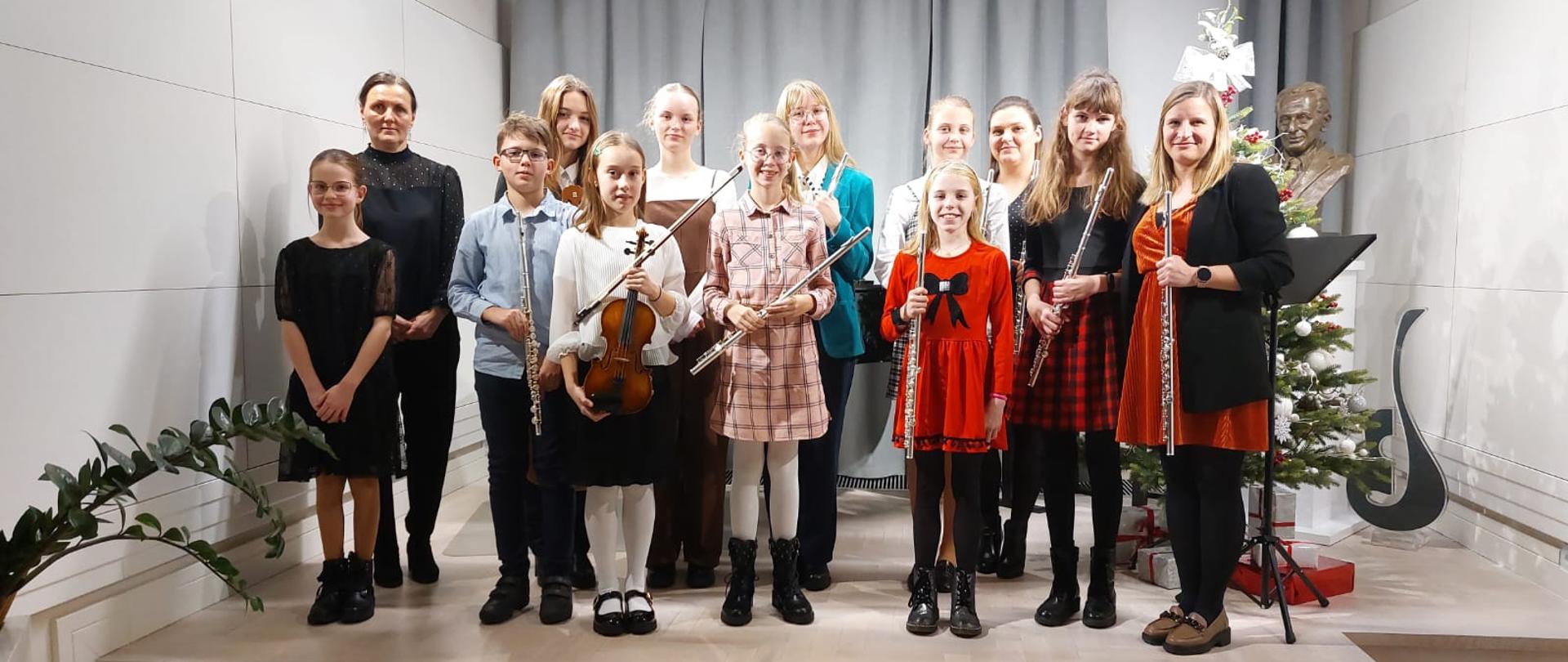 Zdjęcie przedstawia uczniów stojących w dwóch rzędach ubranych na kolorowo. Z prawej strony stroi nauczyciel klasy fletu, a z lewej pani akompaniator. Uczniowie trzymają flety, a jedna uczennica stojąca w pierwszym rzędzie skrzypce. Z prawej strony widać ubraną choinkę, popiersie patrona szkoły i statuetkę logo szkoły. W tle widać zasłonięte częściowo kotary w szarym kolorze. 