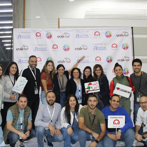 Grupa kobiet i mężczyzn w różnym wieku pozuje na tle roll-upa z logo Polska pomoc, Universite Antonine i Be Baldi