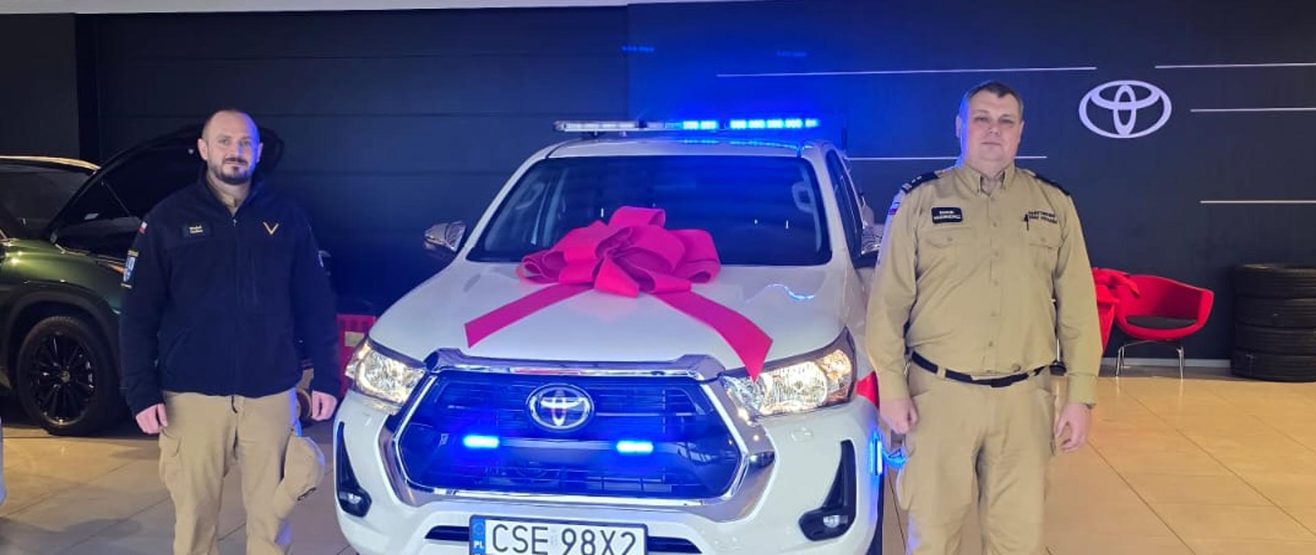 Komendant Powiatowy Państwowej Straży Pożarnej w Sępólnie Krajeńskim w mundurze służbowym po prawej oraz ogniomistrz Michał Delikat po lewej, między nimi samochód biała toyota hilux z dużą czerwoną kokardą na masce. 