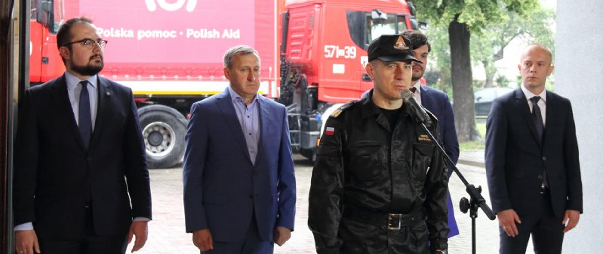 Polscy strażacy ponownie jadą z pomocą – na zdjęciu przemawia nadbryg. Andrzej Bartkowiak komendant główny PSP. W tle ciężarówki z konwoju.