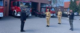 Strażak stoi na baczność komendant główny Państwowej Straży Pożarnej oddaje honor obok stoją pojazdy ratowniczo-gaśnicze.