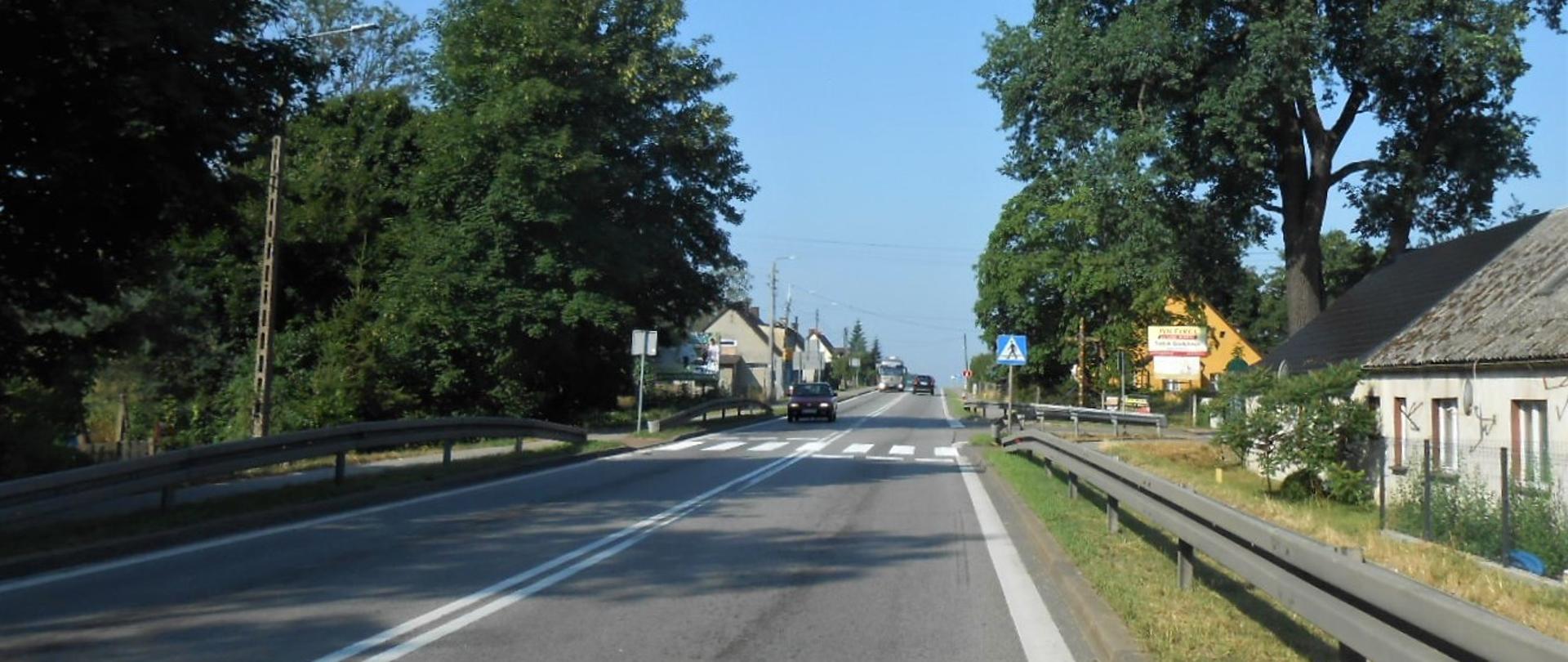 Zdjęcie przedstawia drogę krajową nr 6 w Godętowie w terenie zabudowanym. Na pierwszym planie jest jezdnia z przejściem dla pieszych, a w oddali widać samochody.
