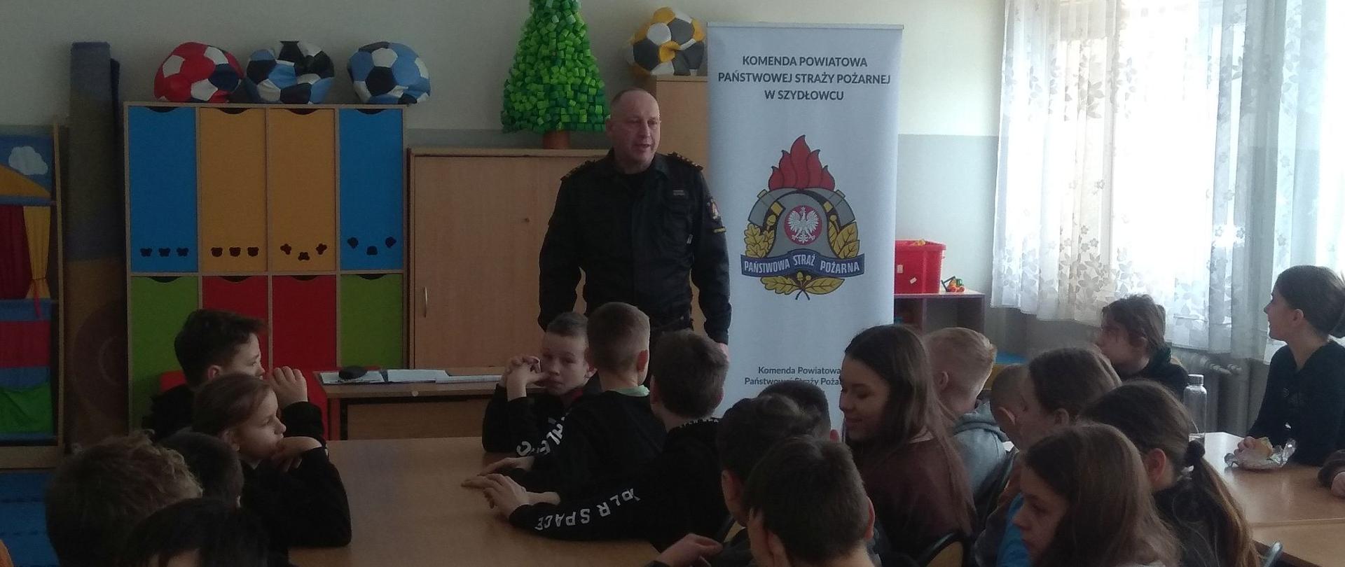 Fotografia przedstawia strażaków podczas prelekcji z dziećmi szkoły podstawowej z terenu powiatu szydłowieckiego na temat bezpieczeństwa podczas ferii zimowych. 