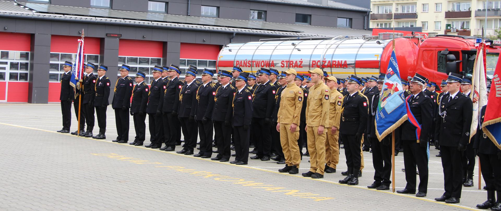 Na placu przed siedzibą Państwowej Straży Pożarnej strażacy oraz poczty sztandarowe uczestniczą w uroczystym apelu z okazji Miejskiego Dnia Strażaka. W tle widoczne są wozy pożarnicze oraz budynek komendy, podkreślające oficjalny charakter wydarzenia.