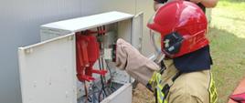Strażak w ubraniu specjalnym i hełmie z opuszczona przyłbicą przy użyciu specjalnej rękawicy wyjmuje ze skrzynki elektrycznej wkładkę bezpiecznikową nożową. W tle widać drugiego strażaka bacznie przyglądającego się wykonywanym czynnościom. 