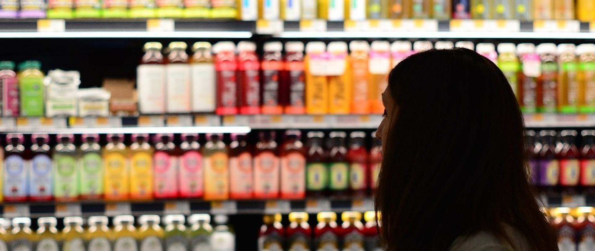 Kobieta w sklepie, patrząca na półki z wieloma kolorowymi napojami.