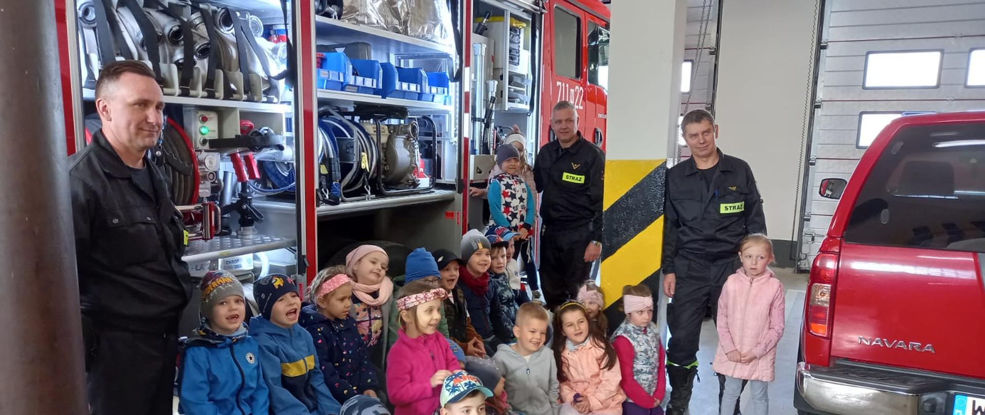 Dzieci z wizytą w Jednostce Ratowniczo – Gaśniczej w Żurominie robią zdjęcie grupowe przy samochodzie gaśniczym ze strażakami.