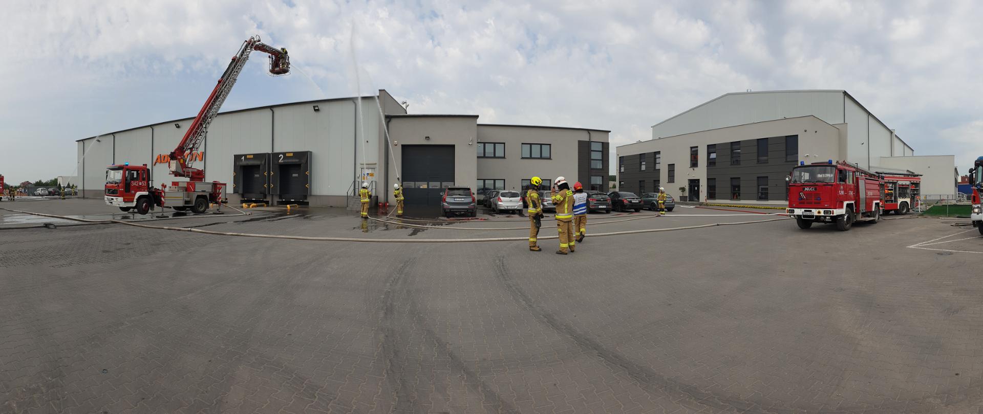 Na zdjęciu widać budynek magazynowy formy Adecon. Na szarej ścianie napis Adecon w kolorze pomarańczowym. Przed magazynem kilka samochodów gaśniczych w kolorze czerwonym z białymi elementami. W centralnym miejscu podnośnik z JRG 2 Kalisz. Strażacy podają dwa prądy wody plus jeden z podnośnika. Ratownicy noszą ubranie specjalne w kolorze piaskowym oraz hełmy. Przed magazynem kostka brukowa w kolorze szarym. Na pierwszym planie widać rozwinięte węże strażackie. Niebo lekko zachmurzone.
