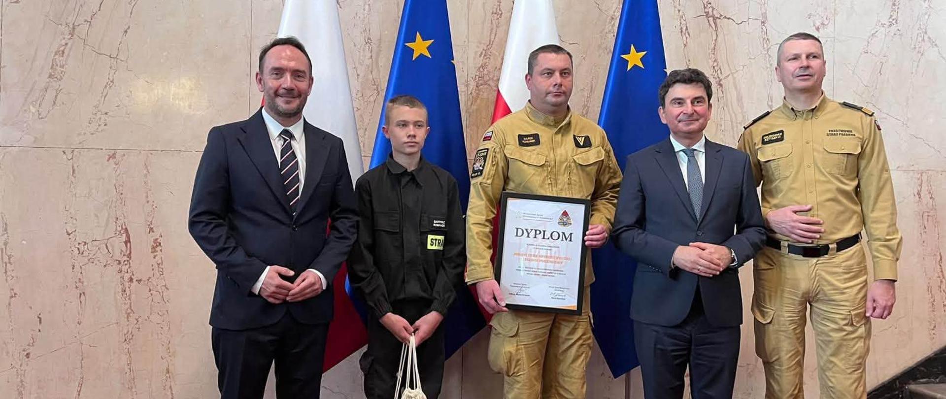 Przedstawiciele Państwowej Straży Pożarnej oraz zaproszeni goście podczas uroczystości wręczenia dyplomu. Na zdjęciu, na tle flag Rzeczypospolitej Polskiej i Unii Europejskiej, pięć osób prezentuje pamiątkowy dyplom. Obok strażaków w umundurowaniu znajdują się dwaj mężczyźni w garniturach oraz młody uczestnik wydarzenia.