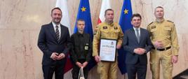 Przedstawiciele Państwowej Straży Pożarnej oraz zaproszeni goście podczas uroczystości wręczenia dyplomu. Na zdjęciu, na tle flag Rzeczypospolitej Polskiej i Unii Europejskiej, pięć osób prezentuje pamiątkowy dyplom. Obok strażaków w umundurowaniu znajdują się dwaj mężczyźni w garniturach oraz młody uczestnik wydarzenia.