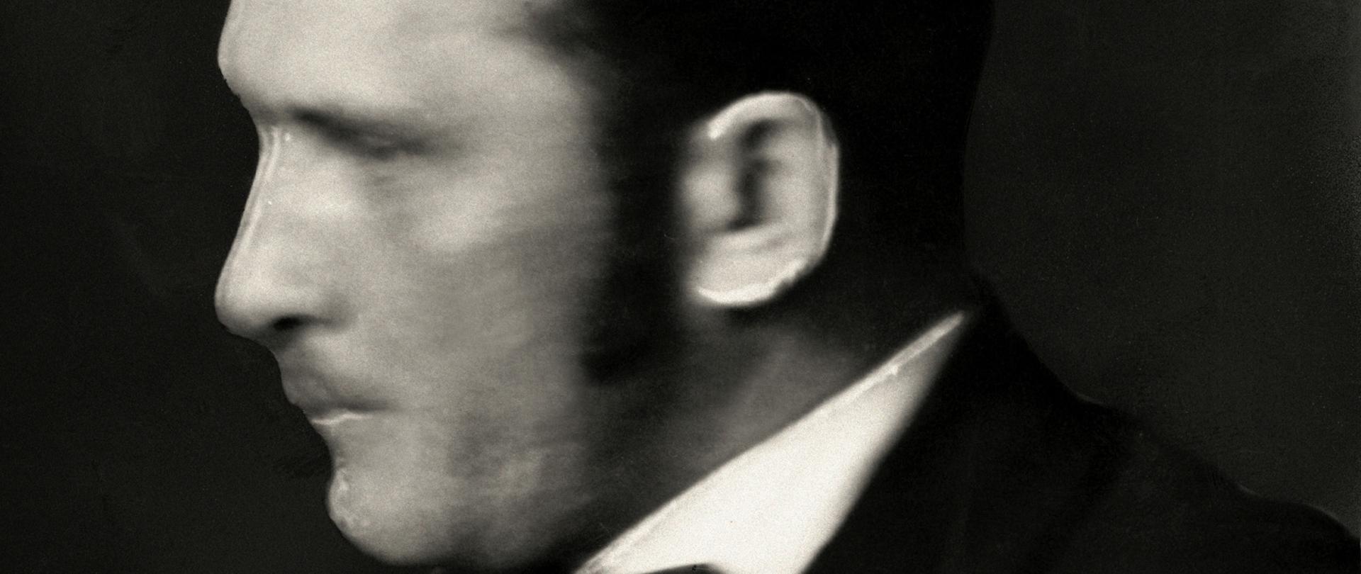 Marian Seyda - czarno-biały portret profilowy mężczyzny z wąsem, ciemne włosy, czarna marynarka, czarna mucha i biała koszula.