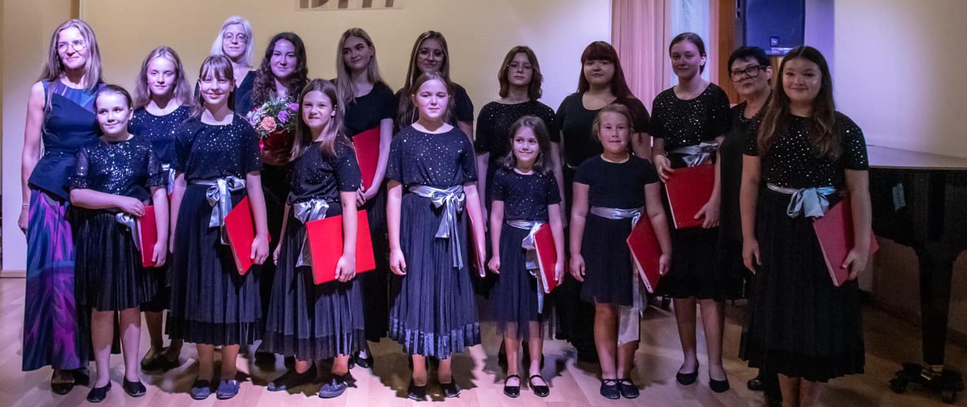 Zdjęcie chóru na scenie w sali koncertowej. Chór ubrany w czarne spódnice, czarne buty, w rękach każdy trzyma czerwoną teczkę. Pani Dyrektor ubrana w mieniącą się odcieniami fioletu spódnicę oraz czarną elegancką bluzkę bez rękawów.