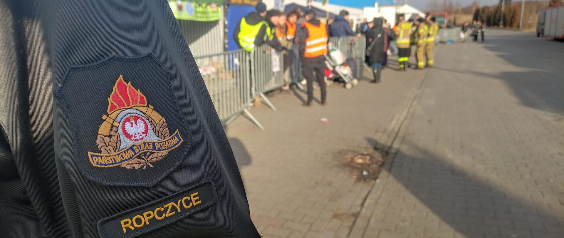 Fragment munduru z naszywką z napisem Ropczyce w tle widok na plac i fragment budynku hali kijowskiej gdzie w tle widać ludzi