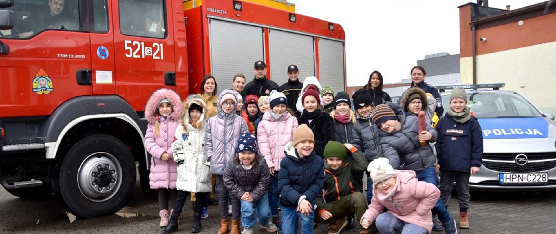 Zdjęcie przedstawia biorące udział w projekcie dzieci wraz z opiekunami. W tle samochód pożarniczy i policyjny, prowadzący wykłady, umundurowani. Dzieci uśmiechnięte, w kurtkach.