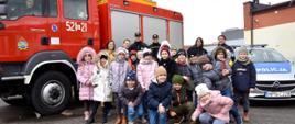 Zdjęcie przedstawia biorące udział w projekcie dzieci wraz z opiekunami. W tle samochód pożarniczy i policyjny, prowadzący wykłady, umundurowani. Dzieci uśmiechnięte, w kurtkach.