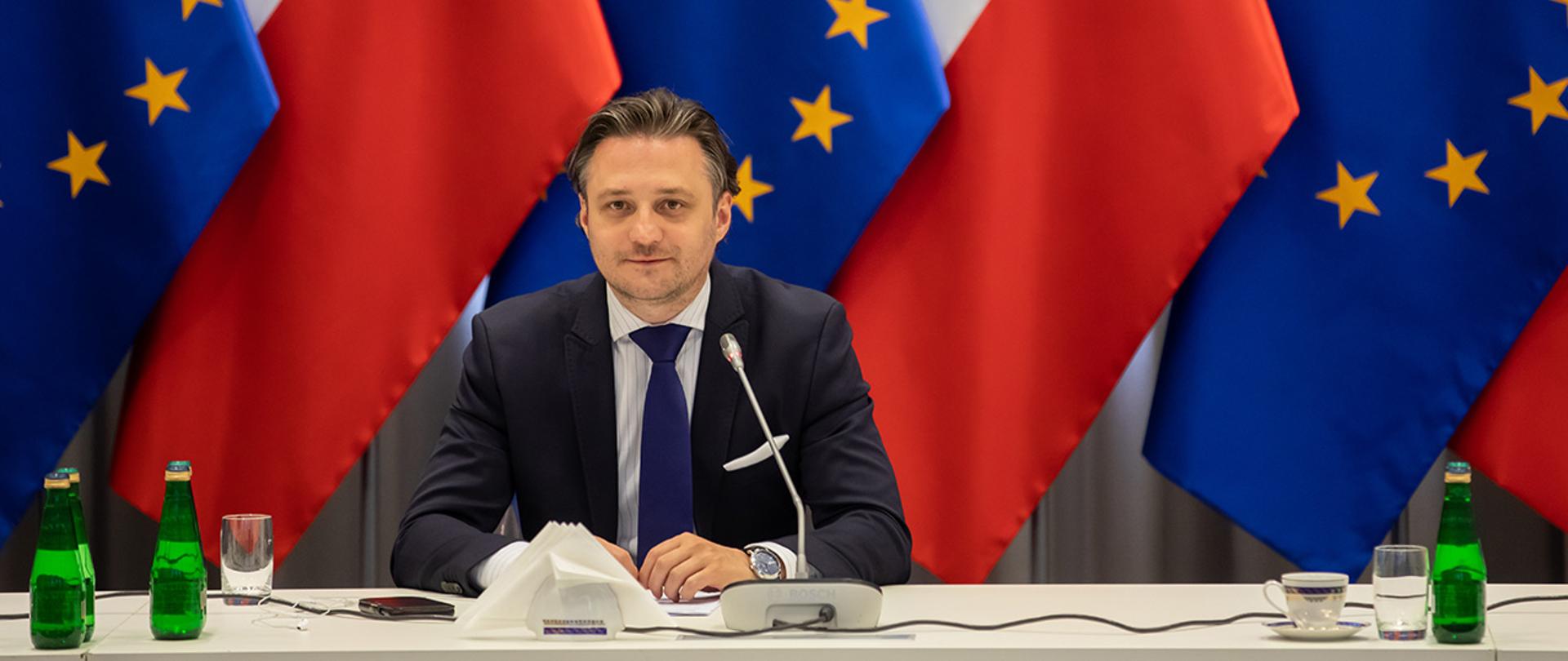 Na zdjęciu widać siedzącego przy stole, uśmiechniętego Bartosza Grodeckiego wiceministra SWiA. W tle widać flagi Polski i UE.