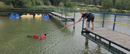 Strażaka za pomocą bosaka teleskopowego próbujący wyciągnąć poszkodowanego który jest w wodzie 