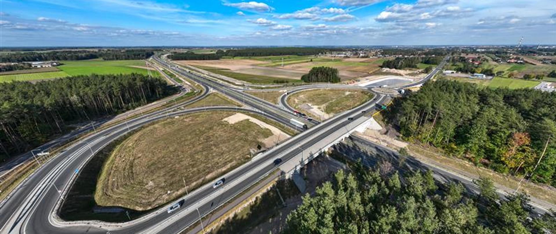 Widok z lotu ptaka na węzeł drogowy w otoczeniu lasów i pól, z kilkoma pasami ruchu, rondami i wiaduktem. Na drogach widoczne są samochody, a w tle rozciąga się krajobraz wiejski z zabudowaniami i niebem z chmurami.