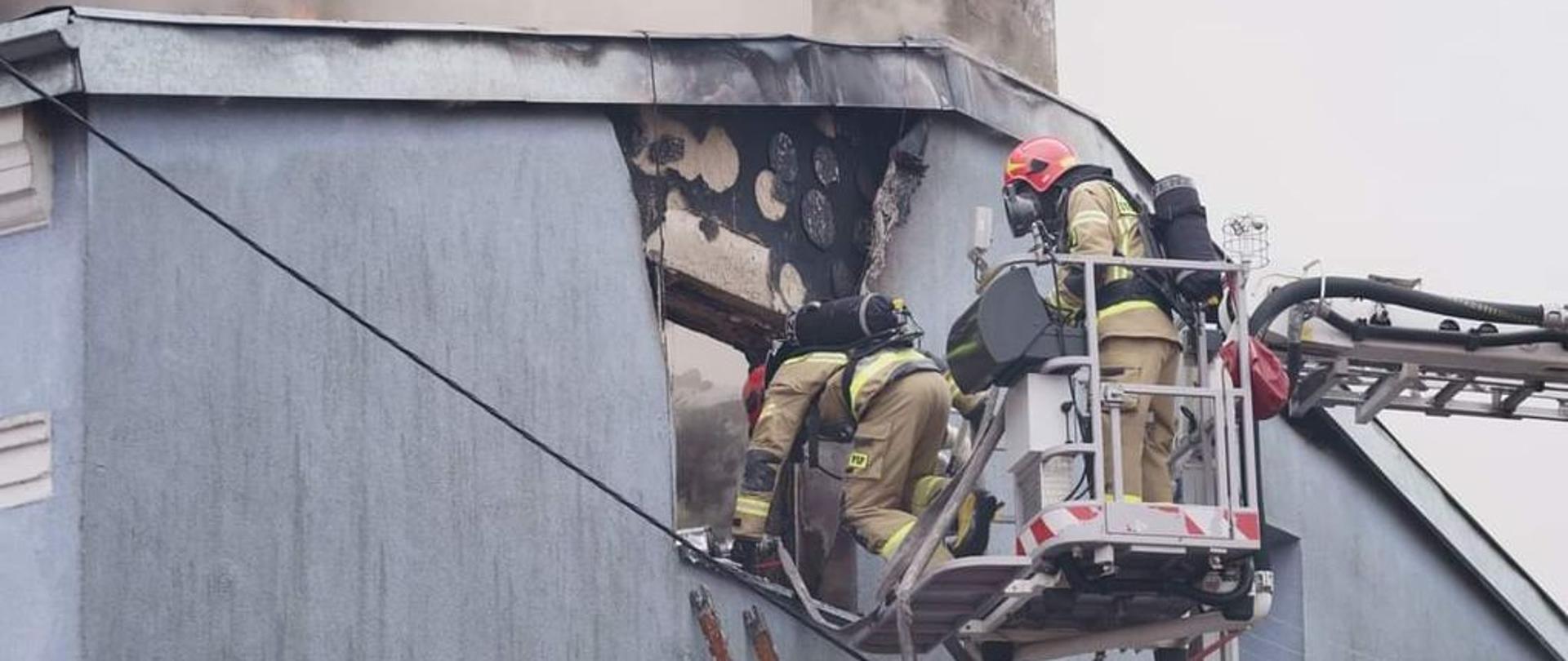 Akcja gaśnicza kamienicy. Strażacy z kosza podnośnika wchodzą przez okno do środka pomieszczenia. Do okna przystawiona jest również drabina. Jest dzień.